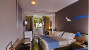 A hotel room with a bed, a desk, a chair, a TV, and a balcony with a view of trees. - Mantenia Hotel (Rethymno)
