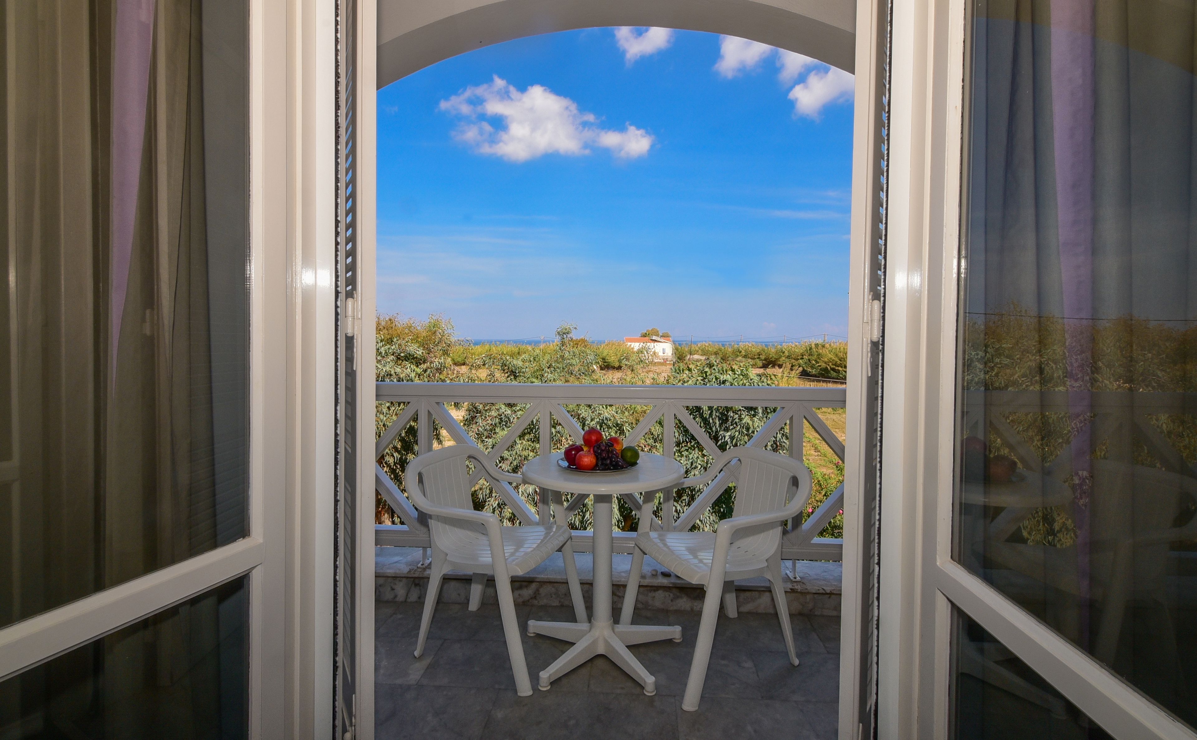 Balkon med hvide stole og et bord med frugt, udover et landskab med grønt og en bygning i det fjerne.