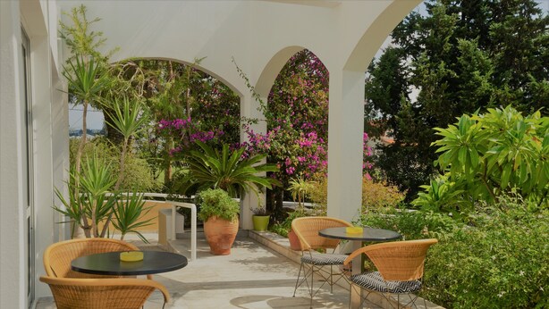An outdoor patio with wicker chairs and a round table, surrounded by potted plants and flowering shrubs. - Mantenia Hotel (Rethymno)