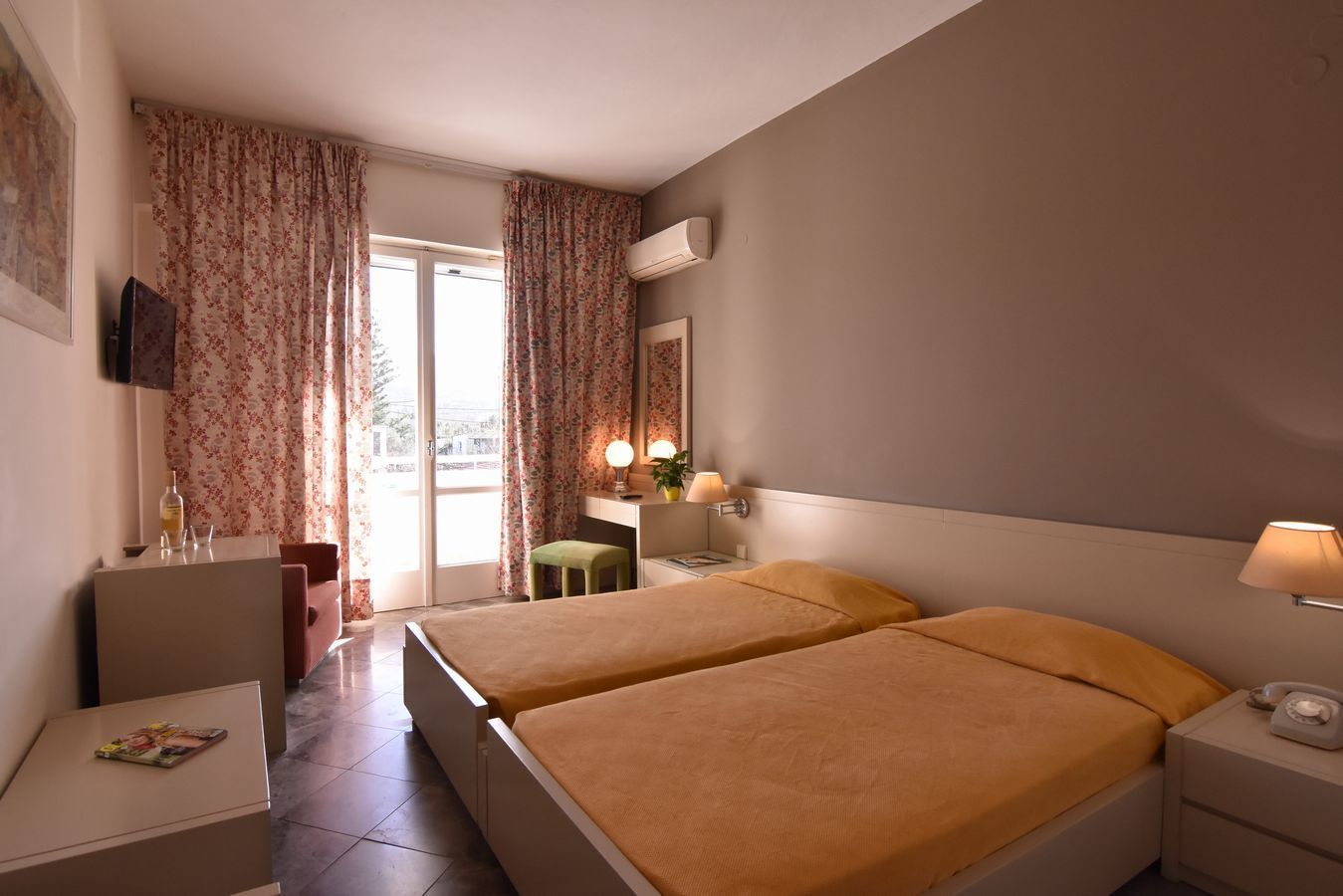 A hotel room with two beds, a desk, a chair, a small table, a telephone, a lamp, a mirror, and a window with curtains.