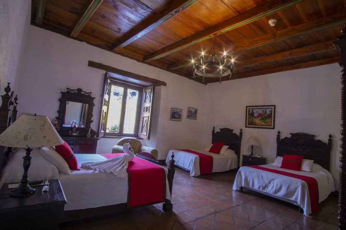 A hotel room with two beds, a chandelier, wooden ceiling beams, a window, and a painting on the wall.