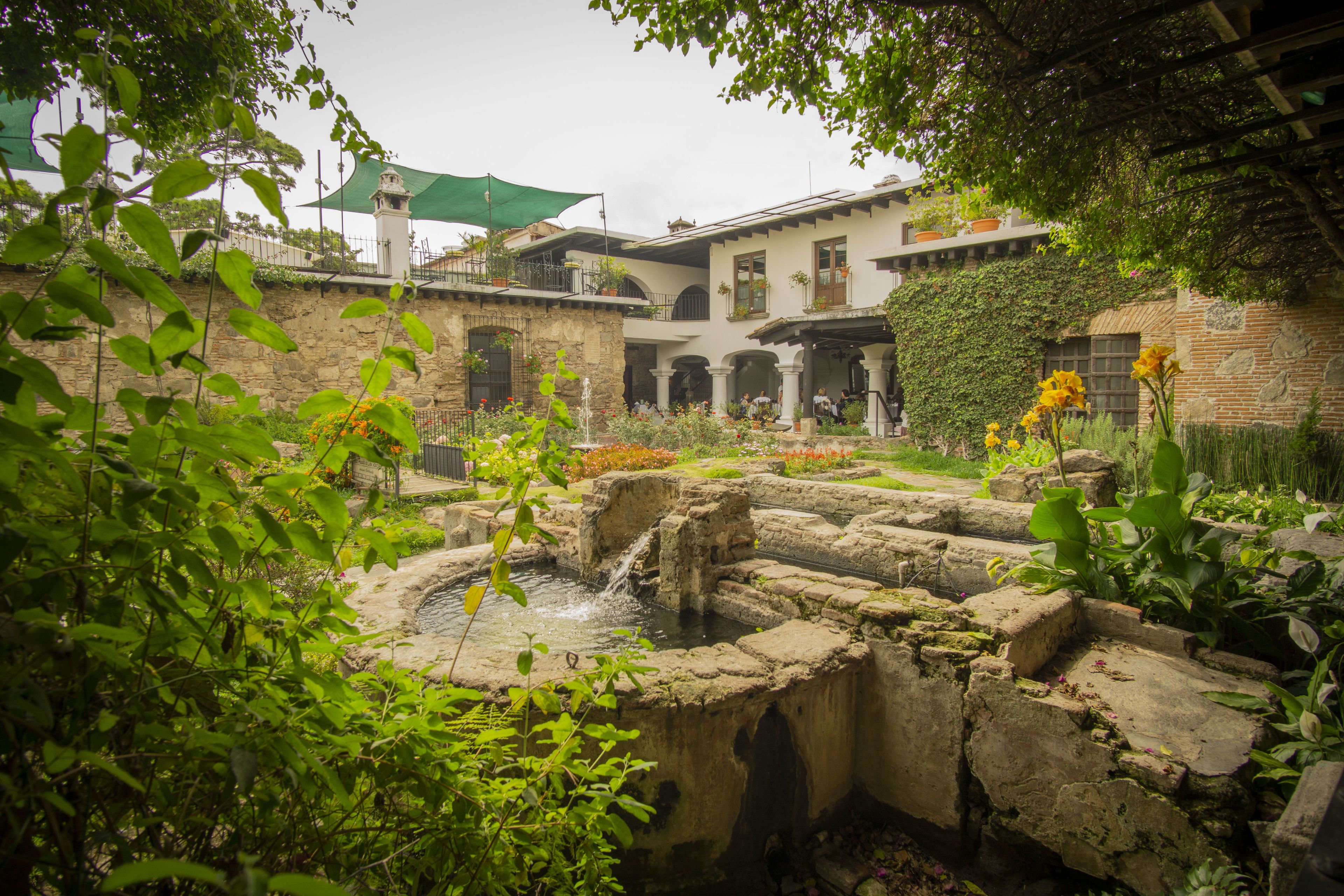 Une cour avec une fontaine en pierre, une végétation luxuriante et un bâtiment doté de nombreux balcons et de plantes suspendues.