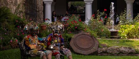 Deux personnes, vêtues de costumes traditionnels, sont assises sur un banc dans un jardin agrémenté d’une fontaine et de plantes en pot.