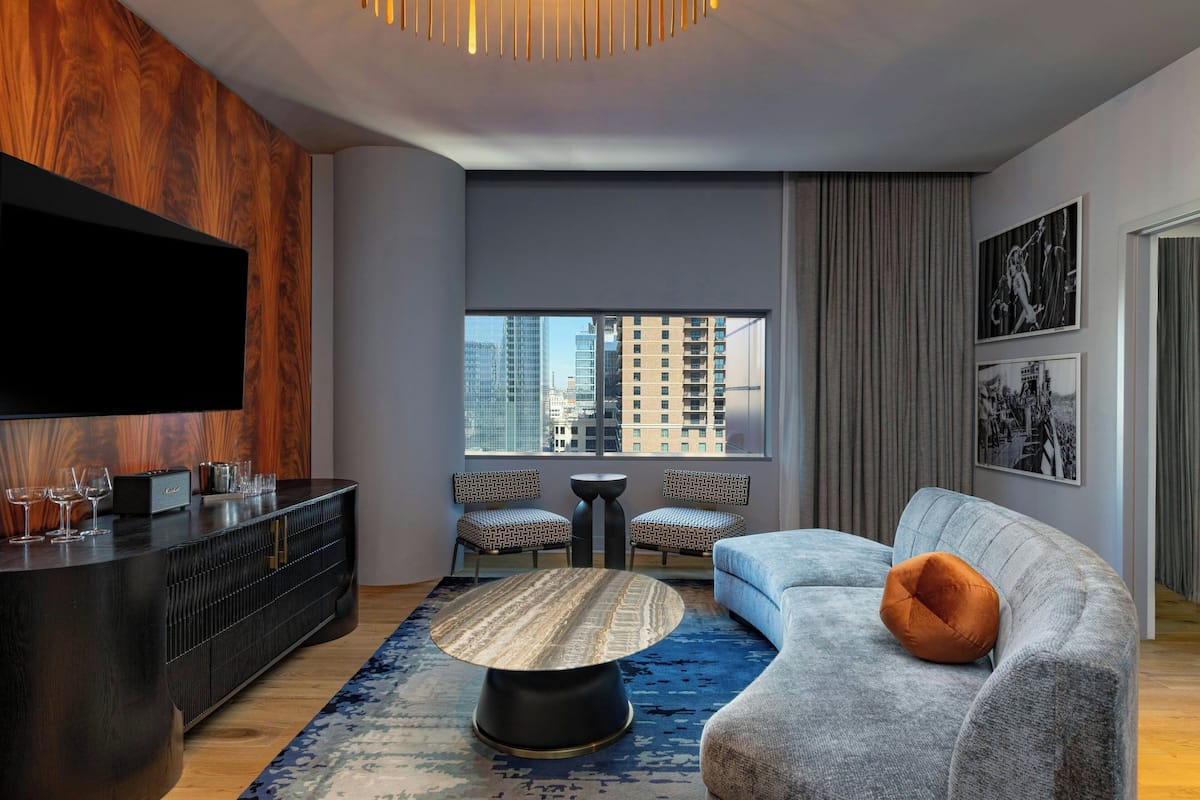 A modern living room with a large flat-screen TV, a dark wooden cabinet, two patterned chairs, a grey sofa with an orange cushion, a round coffee table, and a view of tall buildings through a large window.
