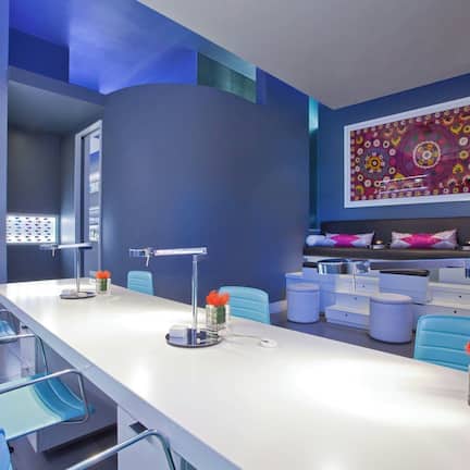A modern hotel lobby with a long reception desk, blue chairs, and a large decorative wall piece.