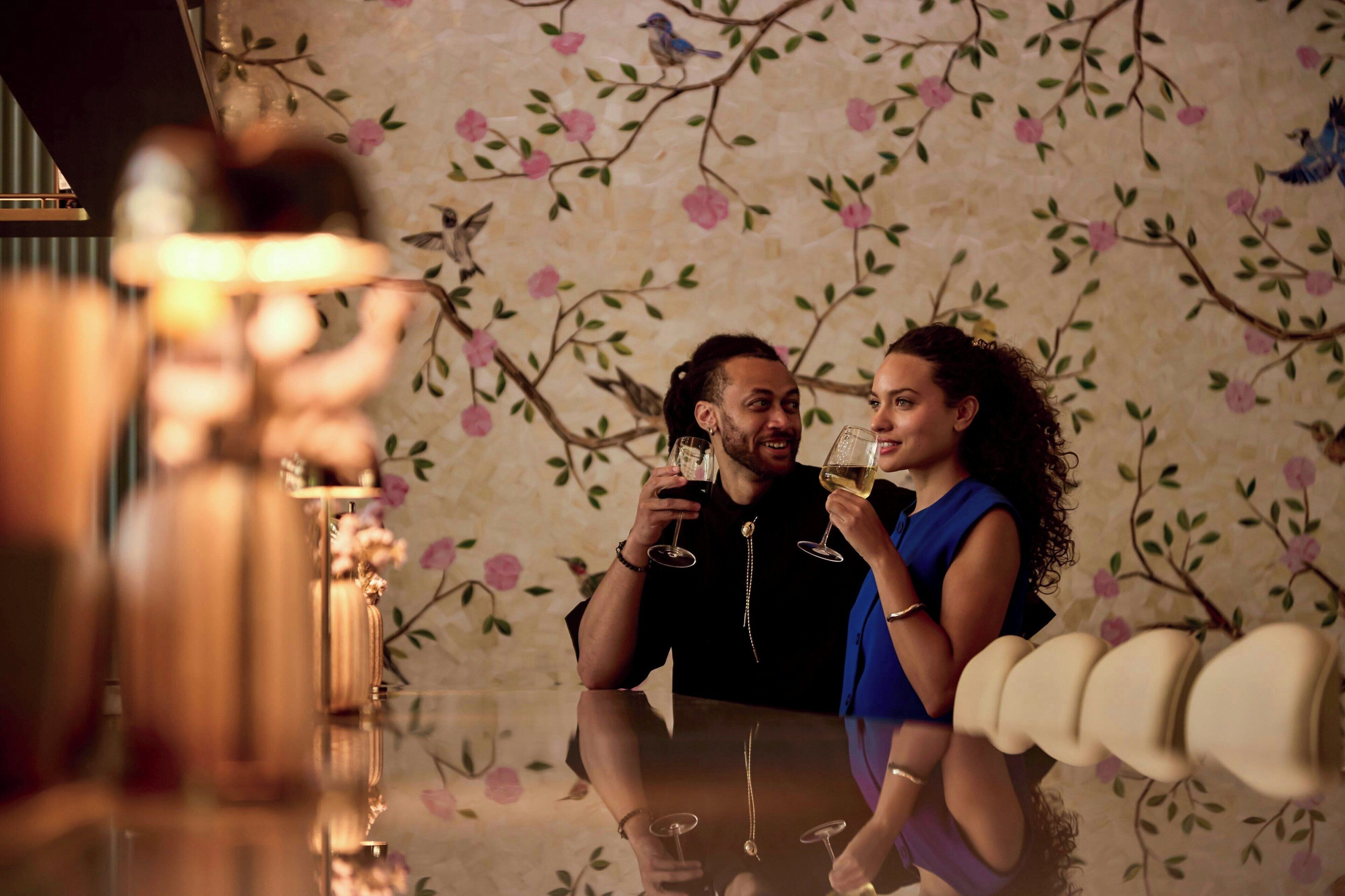 A couple sitting at a table with floral wallpaper, holding wine glasses.