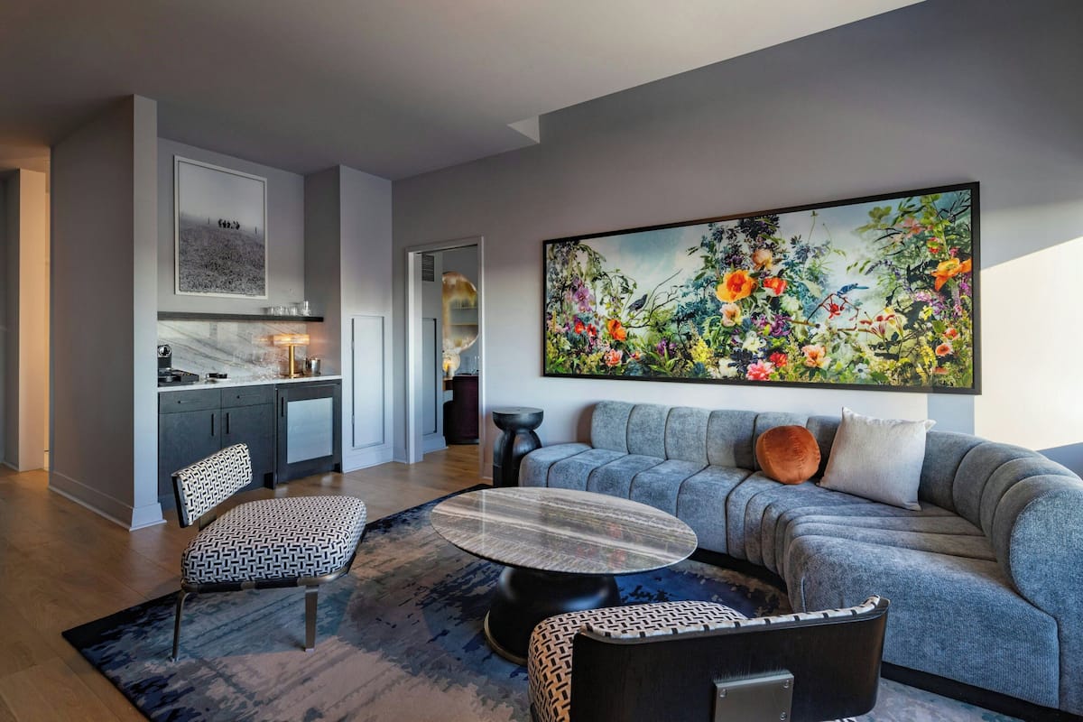 A modern living room with a grey sofa, patterned chairs, a round coffee table, and a large floral painting on the wall.