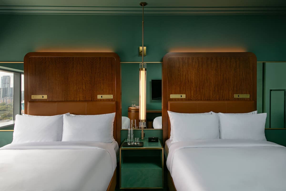 A hotel room with two beds, a nightstand, a lamp, and a view of the city.