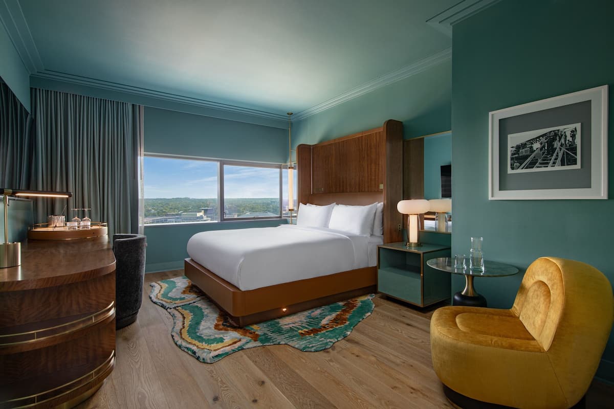 A modern hotel room with a large bed, a wooden desk, a yellow armchair, and a view of the city.