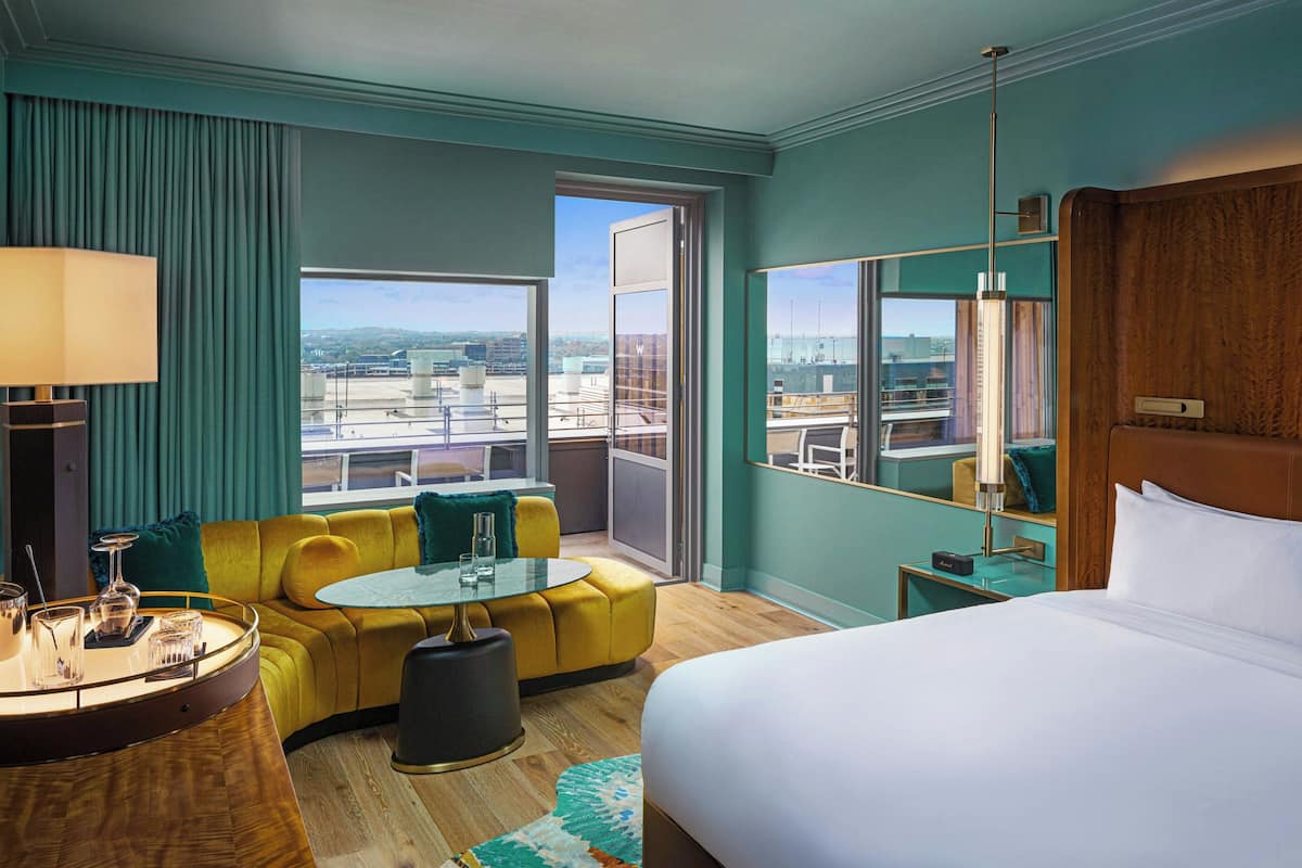 A hotel room with a bed, a sofa, a round table, and a view of the city through large windows.
