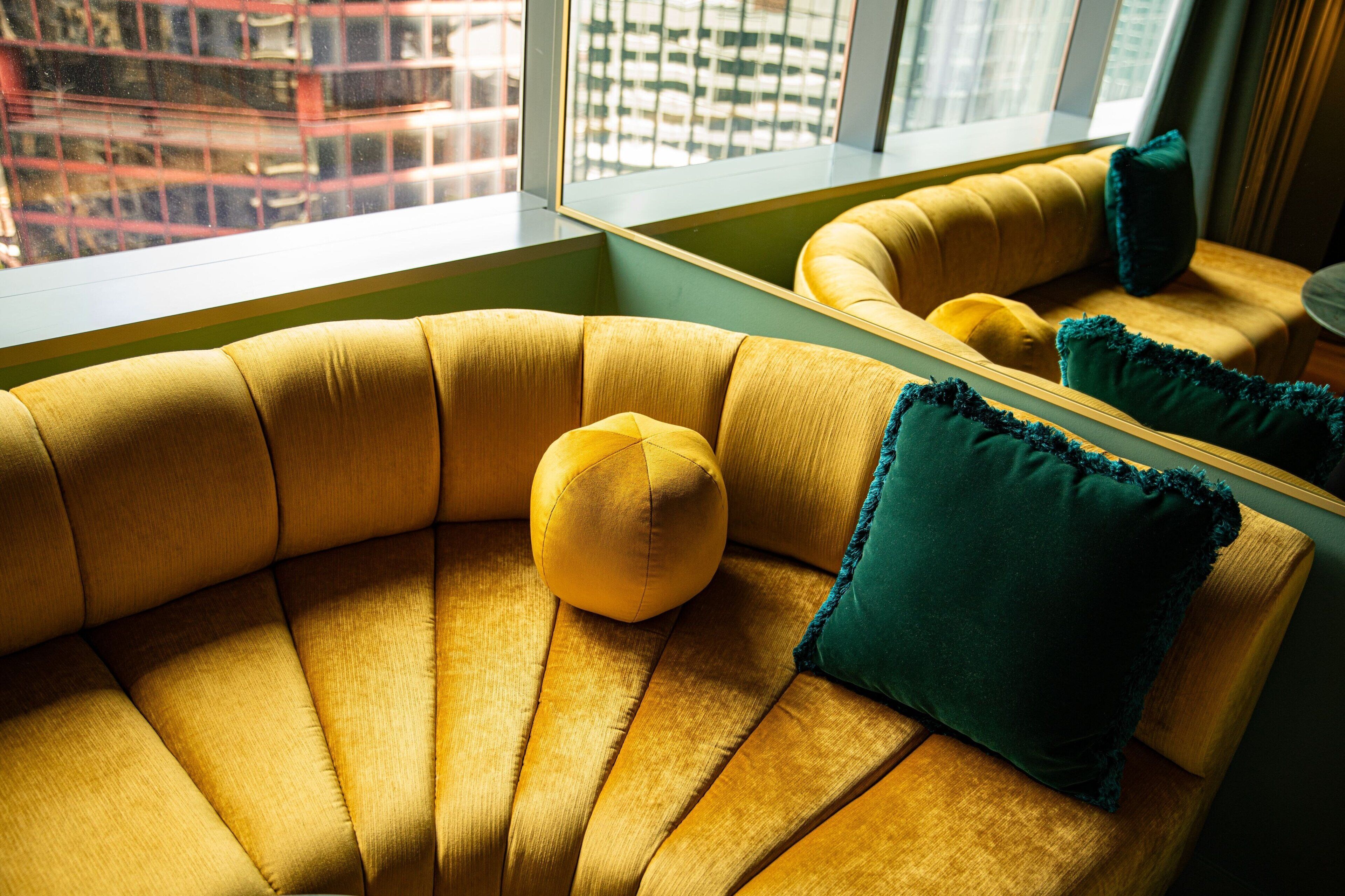 Un sofá curvo con un cojín amarillo y una almohada verde, ubicado cerca de una ventana con vistas a edificios.
