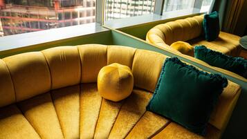 Un sofá curvo con un cojín amarillo y una almohada verde, ubicado cerca de una ventana con vistas a edificios.