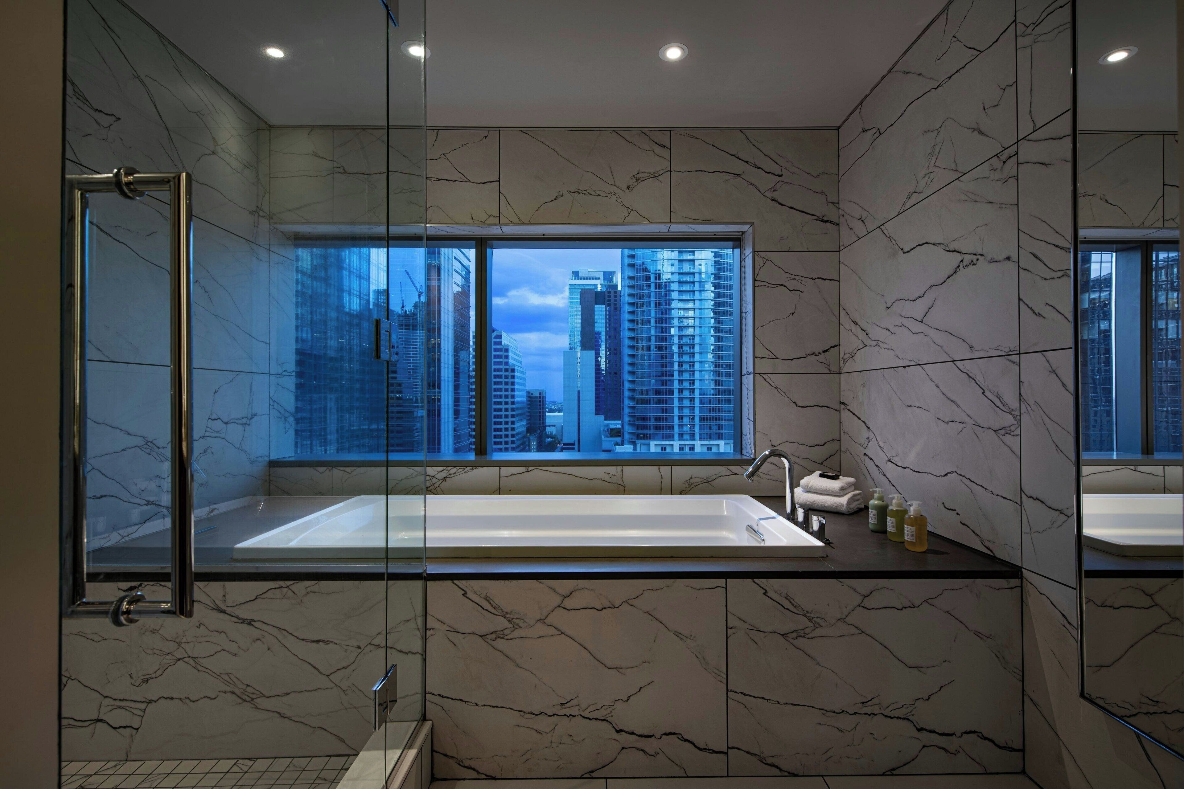 Un baño moderno con una bañera grande, una ducha con mampara de vidrio y vistas a la ciudad a través de un ventanal.
