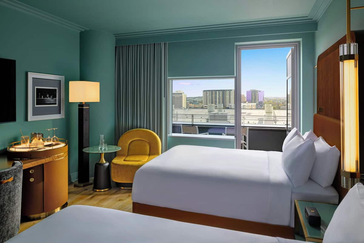 A hotel room with two beds, a desk with a lamp, a chair, and a view of the cityscape through a large window.