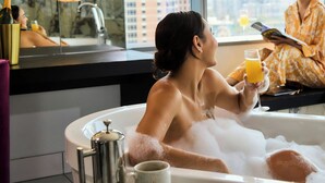 A woman in a bathtub with bubbles, holding a glass of orange juice, and a coffee cup on a tray. - W Austin (Austin)