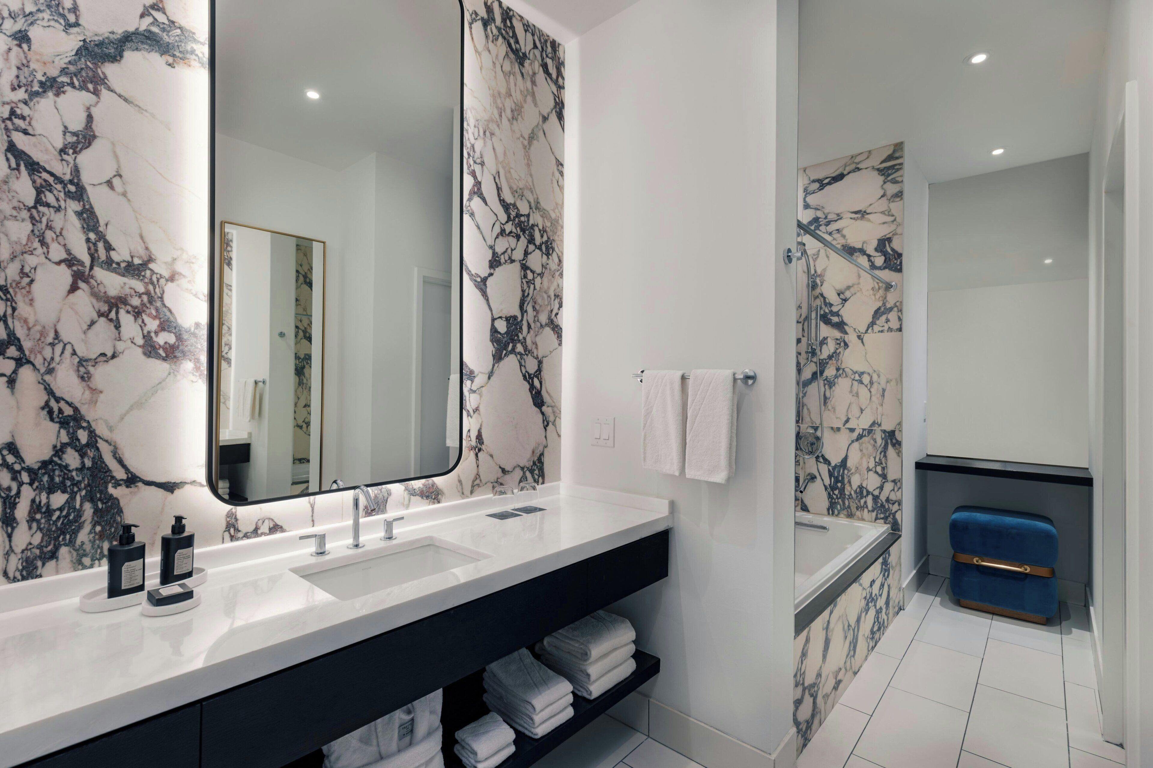 A modern bathroom with a marble backsplash, a large mirror, a white sink, and a blue suitcase.