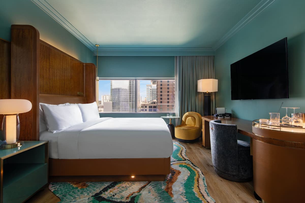 A hotel room with a large bed, a desk with a lamp, a chair, a TV, and a view of the city.
