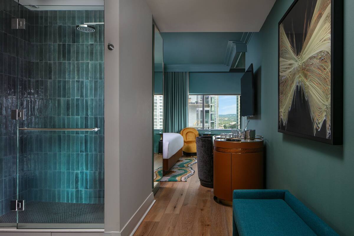 A modern hotel room with a glass-enclosed shower, a bed with a view, a seating area, and a mounted TV.