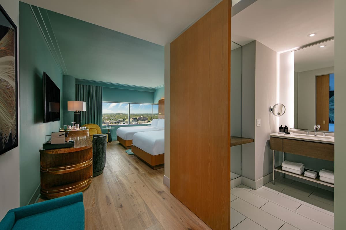 A modern hotel room with a large bed, a wooden side table, a desk with a lamp, a large window with a view, and a bathroom with a mirror and sink.