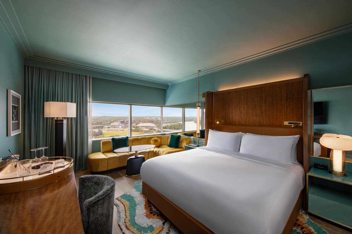 A hotel room with a large bed, a desk with a lamp, a sofa, and a view of the city.