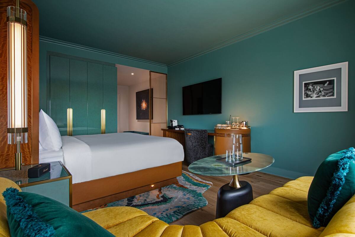 A modern hotel room with a large bed, a sitting area with a yellow sofa, a glass coffee table, and a desk with a computer.
