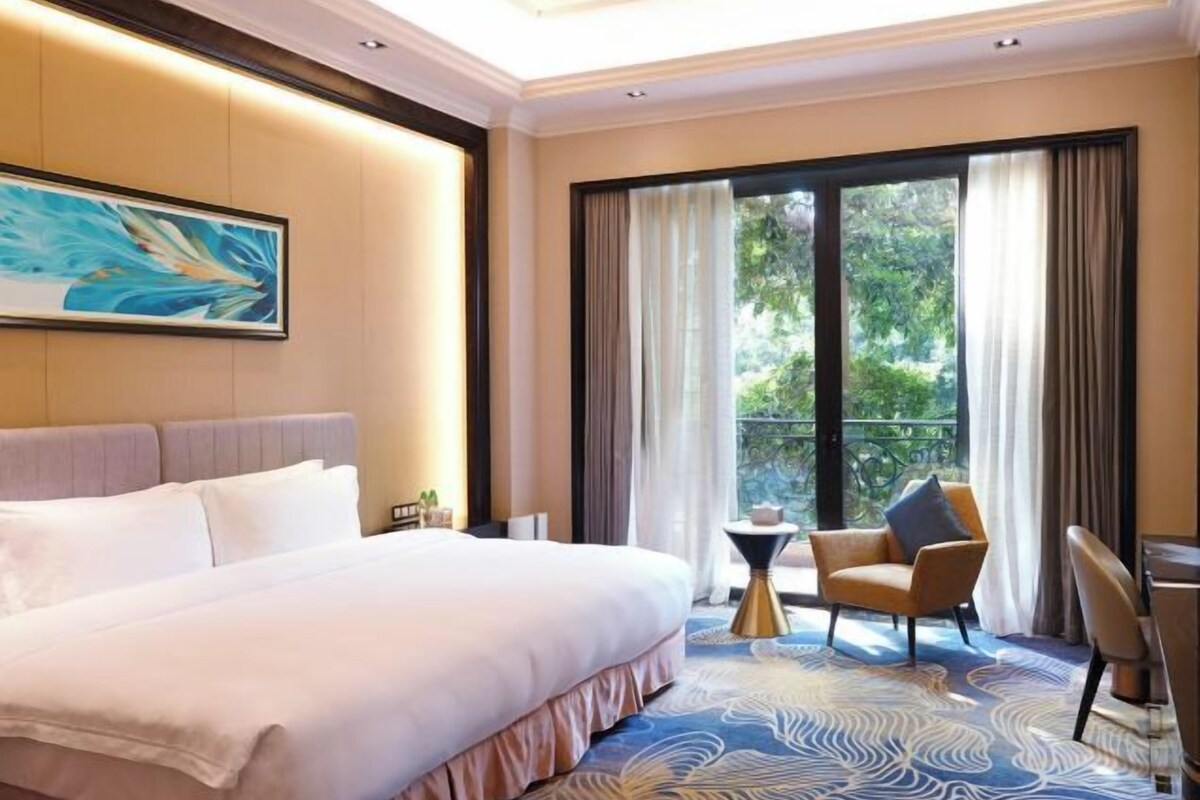 Ein Hotelzimmer mit einem großen Bett, einem kleinen runden Tisch, zwei Stühlen, einem Bild an der Wand und einem Balkon mit Vorhängen.