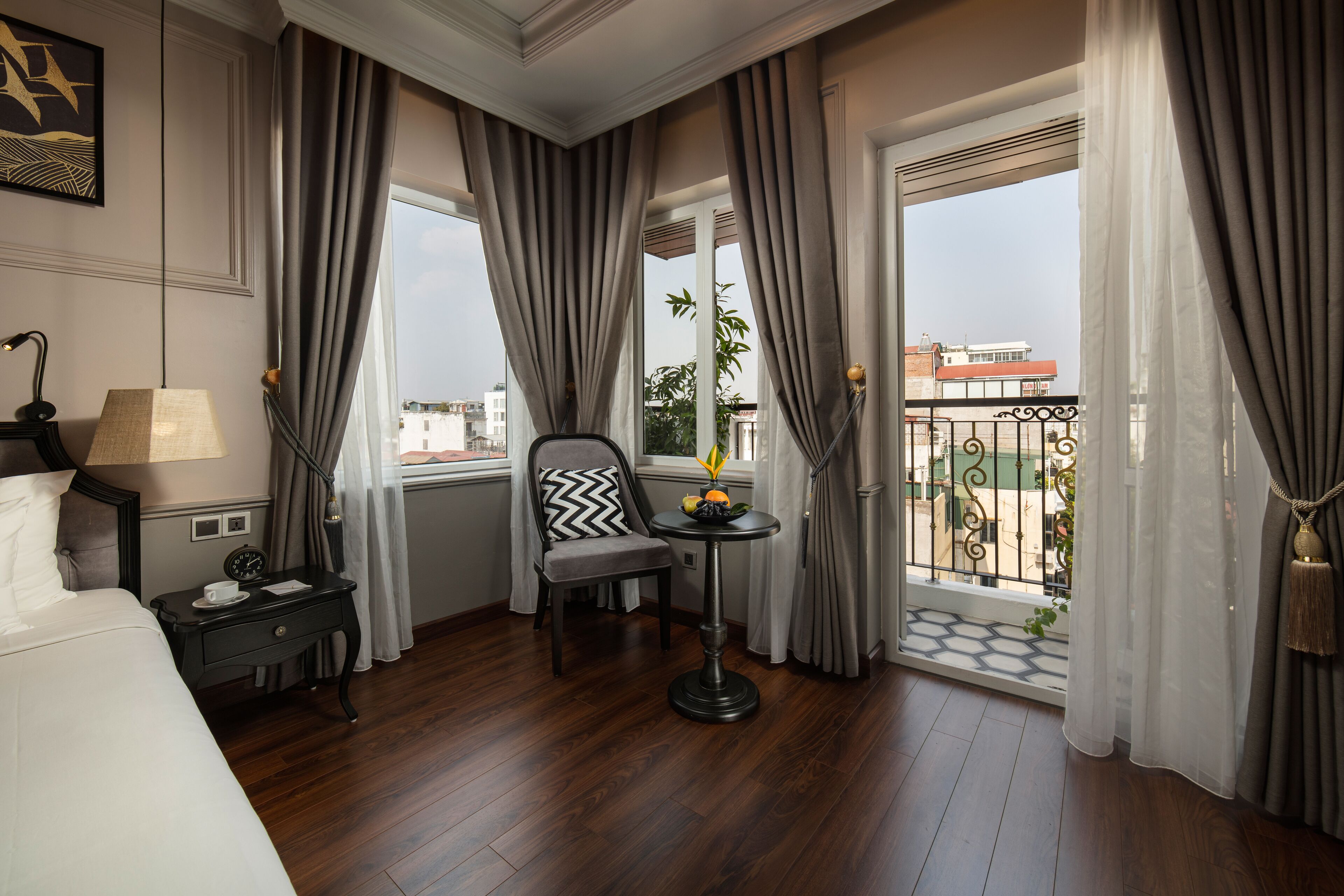 Ein Hotelzimmer mit Bett, Stuhl, kleinem Tisch mit Obst und Balkon mit Blick auf Gebäude und eine Ziegelmauer.