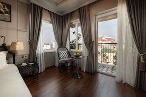 Habitación de hotel con cama, una silla, una mesita con fruta y un balcón con vista a edificios y una valla.