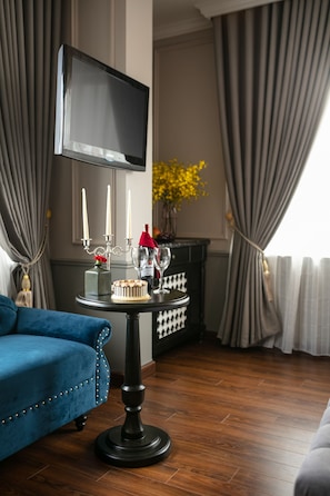 A modern living room with a flat-screen TV mounted on the wall, a round table with candles, a cake, and a vase of yellow flowers. - Imperial Hotel & Spa (Hanoi)
