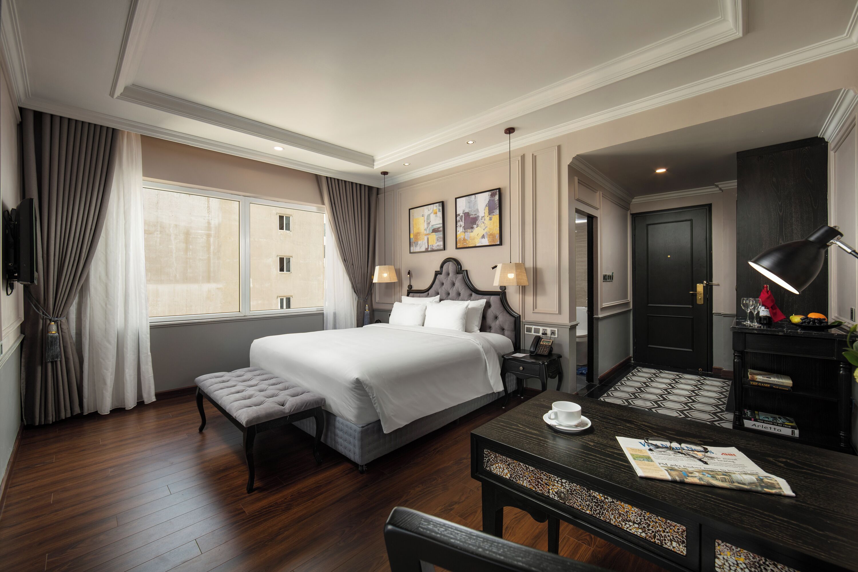 A hotel room with a large bed, a desk with a chair, a small table with a cup and a menu, a television, and a door.