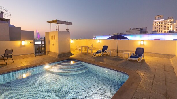 A rooftop pool area with lounge chairs and a small table, surrounded by buildings and a clear sky.