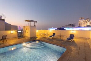Un espace piscine aménagé sur le toit, avec des chaises longues et une petite table, entouré d’immeubles et surplombant un ciel dégagé.