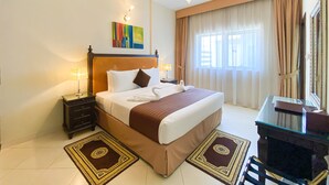 A hotel room with a large bed, a desk with a chair, a desk lamp, a nightstand, a wall-mounted lamp, and a window with curtains.