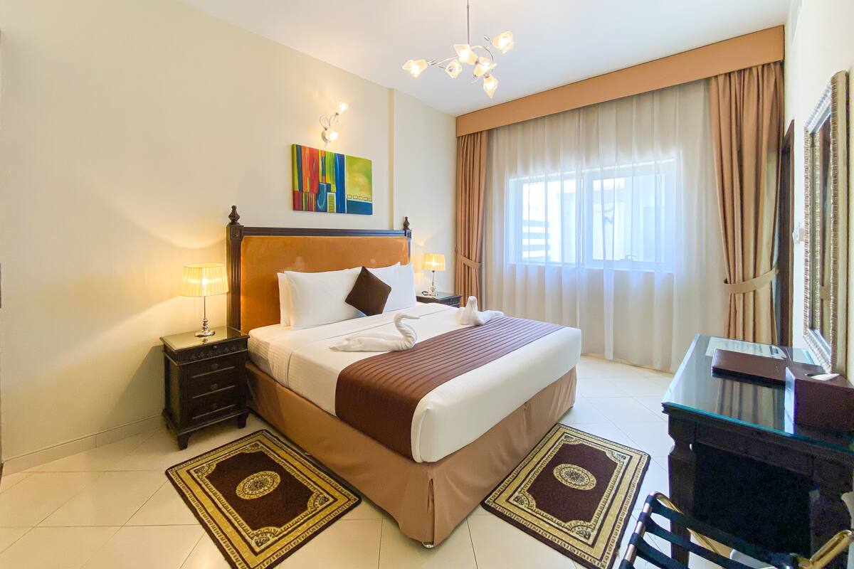 A hotel room with a large bed, a desk with a chair, a desk lamp, a nightstand, a wall-mounted lamp, and a window with curtains.