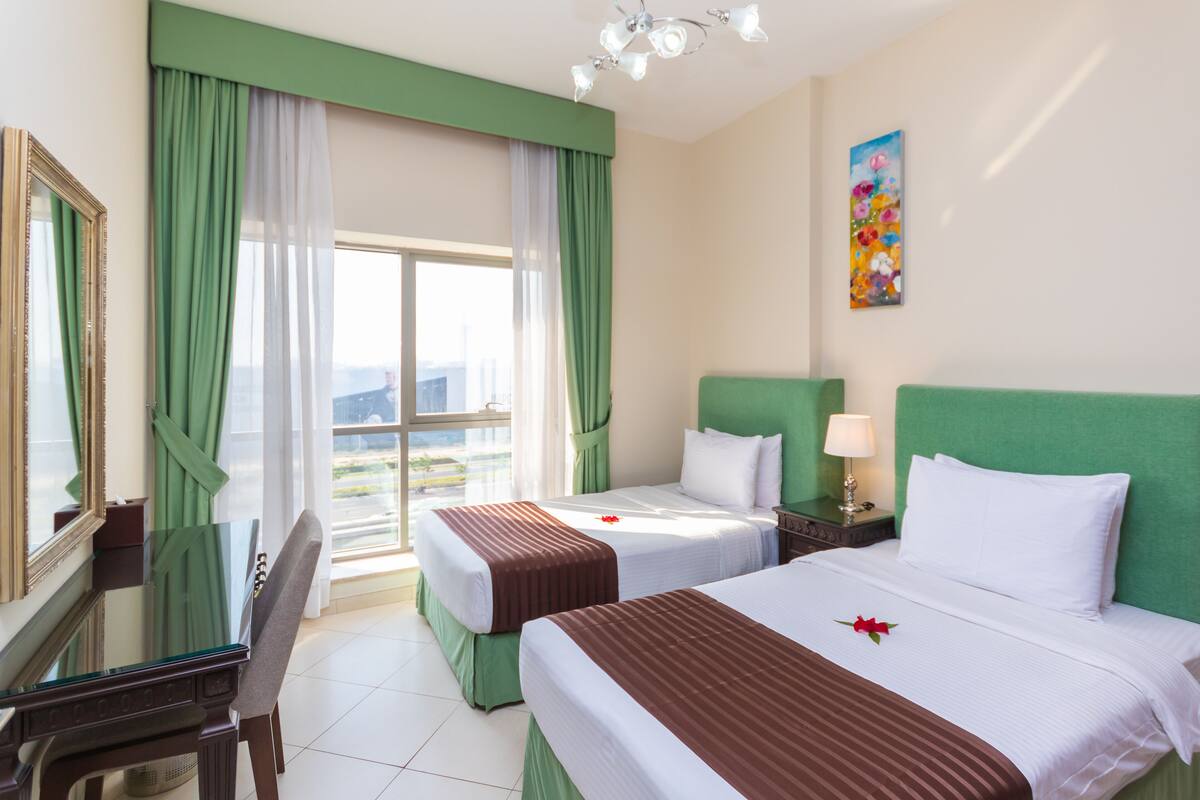 A hotel room with two beds, a desk, a chair, a painting on the wall, and a view of the city through the window.