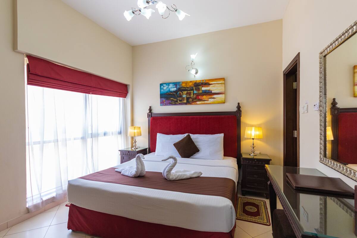 A hotel room with a large bed, a painting on the wall, a mirror, and a window with curtains.