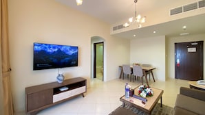A modern living room with a flat-screen TV, a wooden entertainment unit, a dining area with a table and chairs, and a hallway leading to a door.