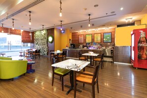 Un restaurant avec un plancher en bois, des tables entourées de chaises vertes et oranges, un réfrigérateur Coca-Cola et un mur orné d’un motif floral.