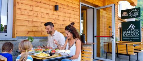 A family dining at an outdoor table with a wooden exterior and a sign for Albergo Dimaro.
