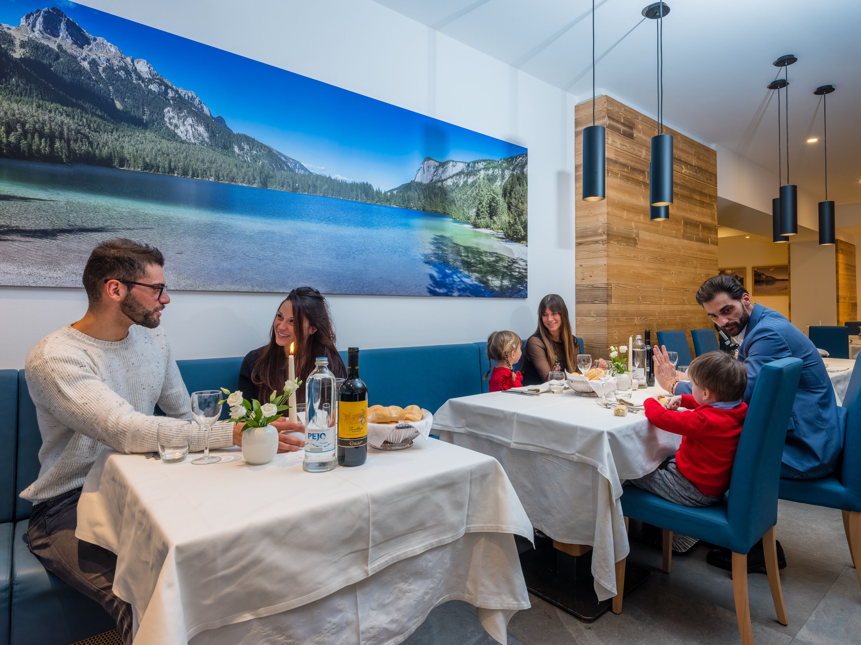 Ein Restaurant mit gedeckten Tischen, einem großen Landschaftsbild von Bergen an der Wand und moderner Pendelleuchten.