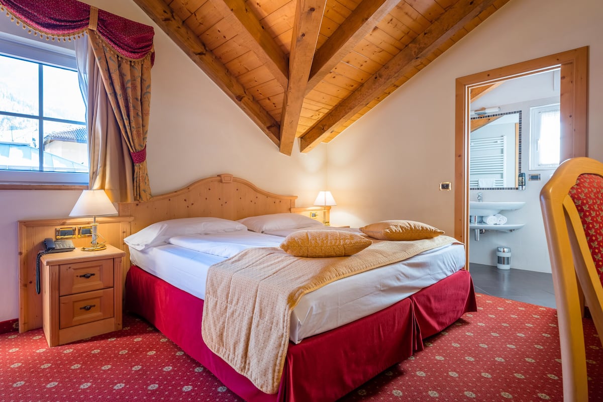 A bedroom with a wooden ceiling, a bed with white linens, a red carpet, and a nightstand with a lamp.