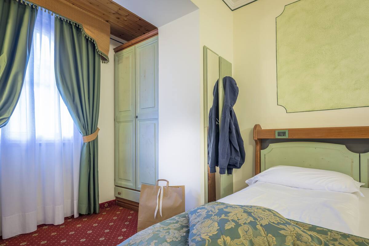 A hotel room with a bed, a wooden headboard, a green patterned pillow, a red carpet, a wooden wardrobe, and a coat hanging on the wall.
