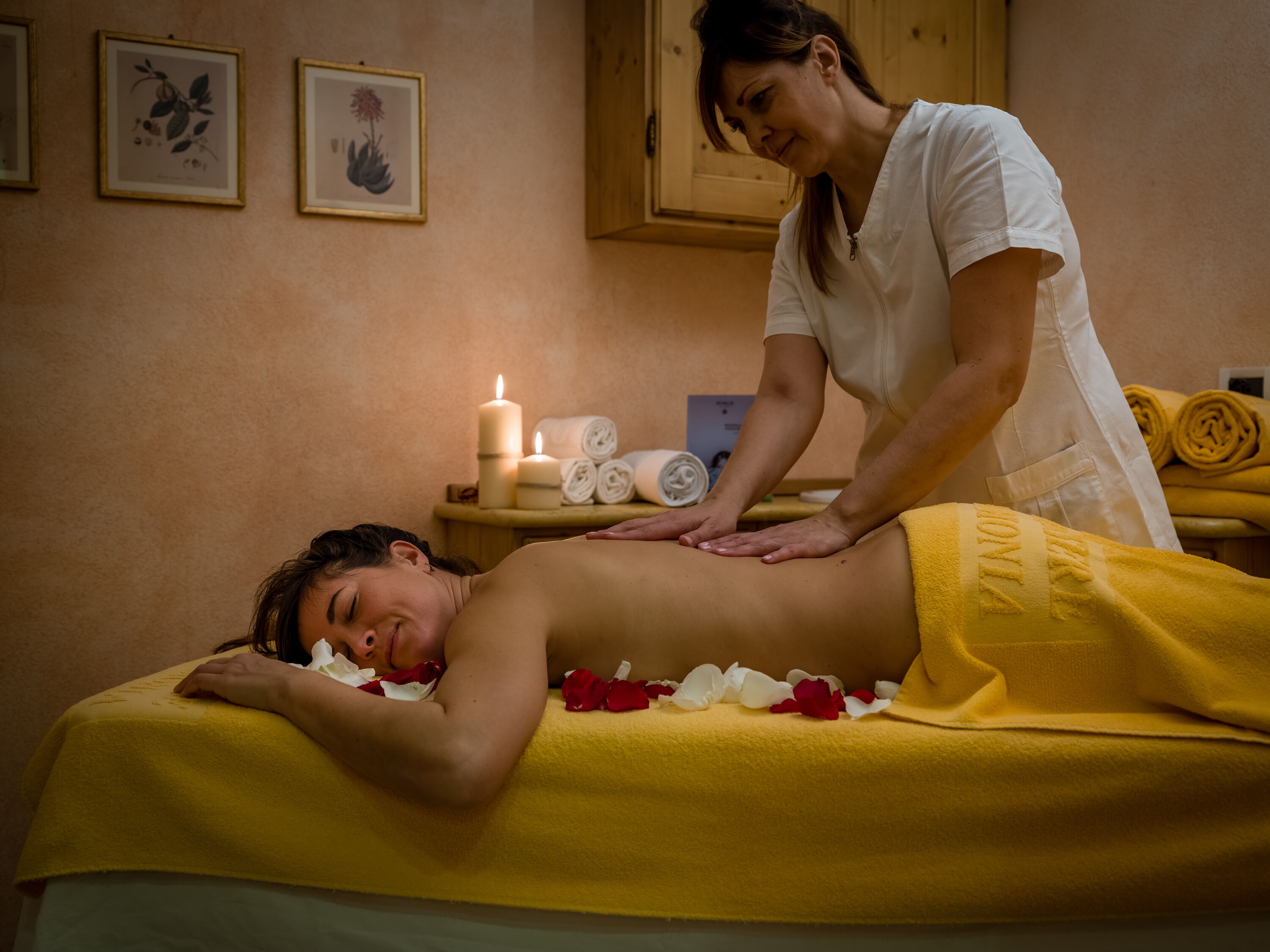 A massage therapy session in a room with a wooden cabinet, framed botanical prints, candles, and towels.