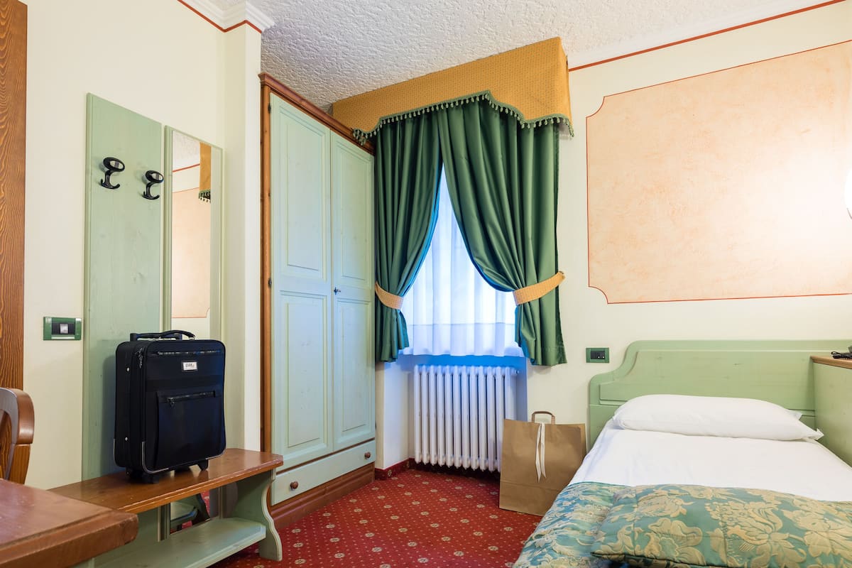 A hotel room with a bed, a desk, a suitcase, a radiator, and a window with curtains.