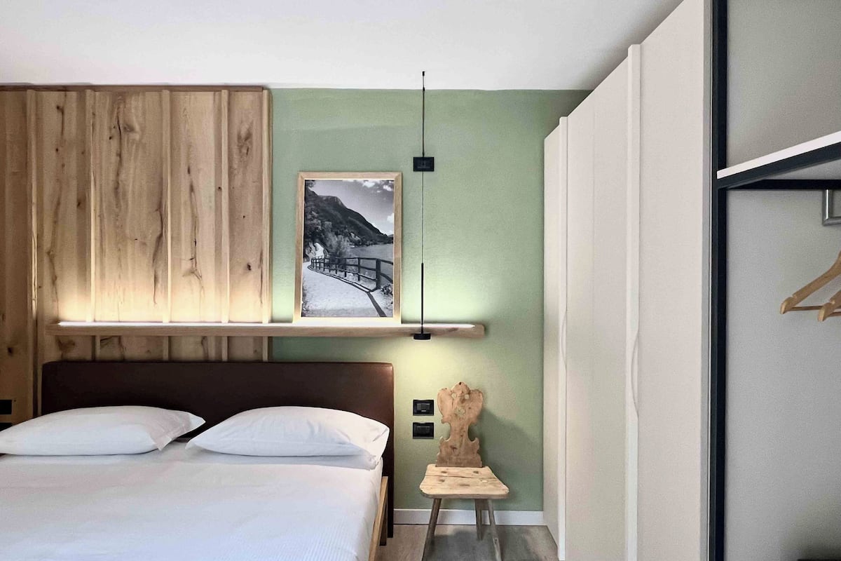 A bedroom with a bed, a wooden headboard, a framed picture, a small wooden stool, and a hanging light fixture.