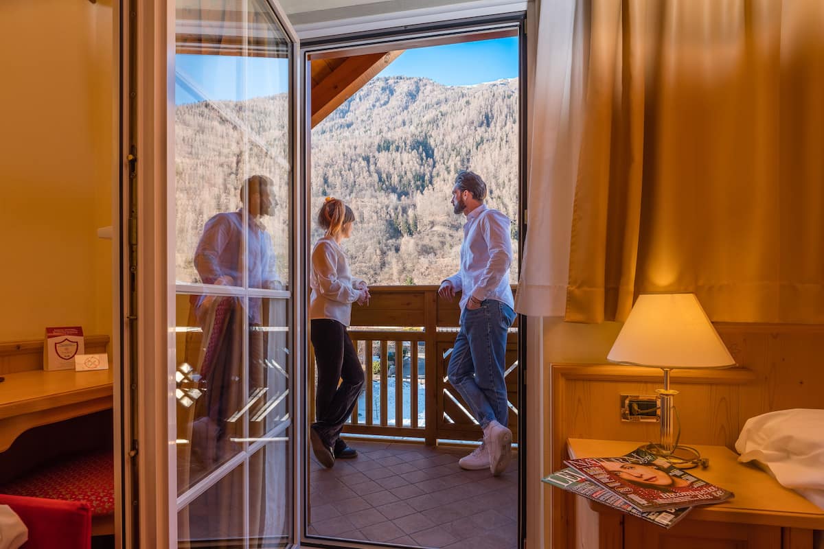 A hotel room with a wooden balcony, a red carpet, a wooden nightstand with a lamp, and a bed with white linens.