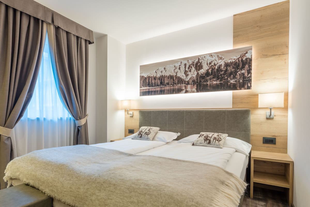 A hotel room with a large bed, two bedside tables, a mounted artwork of a mountain landscape, and a window with curtains.
