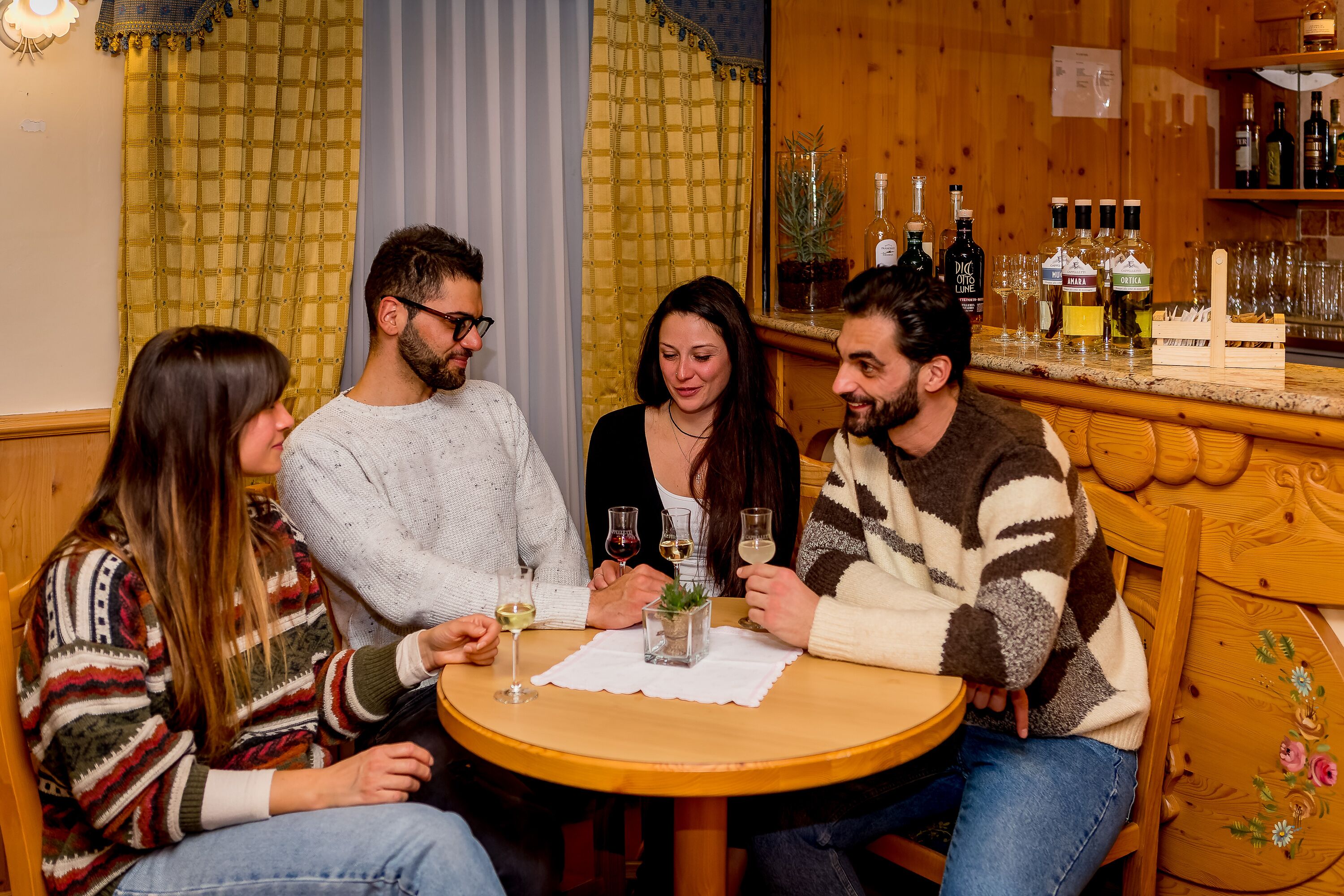 A group of people seated around a wooden table with a white tablecloth, holding wine glasses. Behind them is a wooden bar with various bottles and glasses.