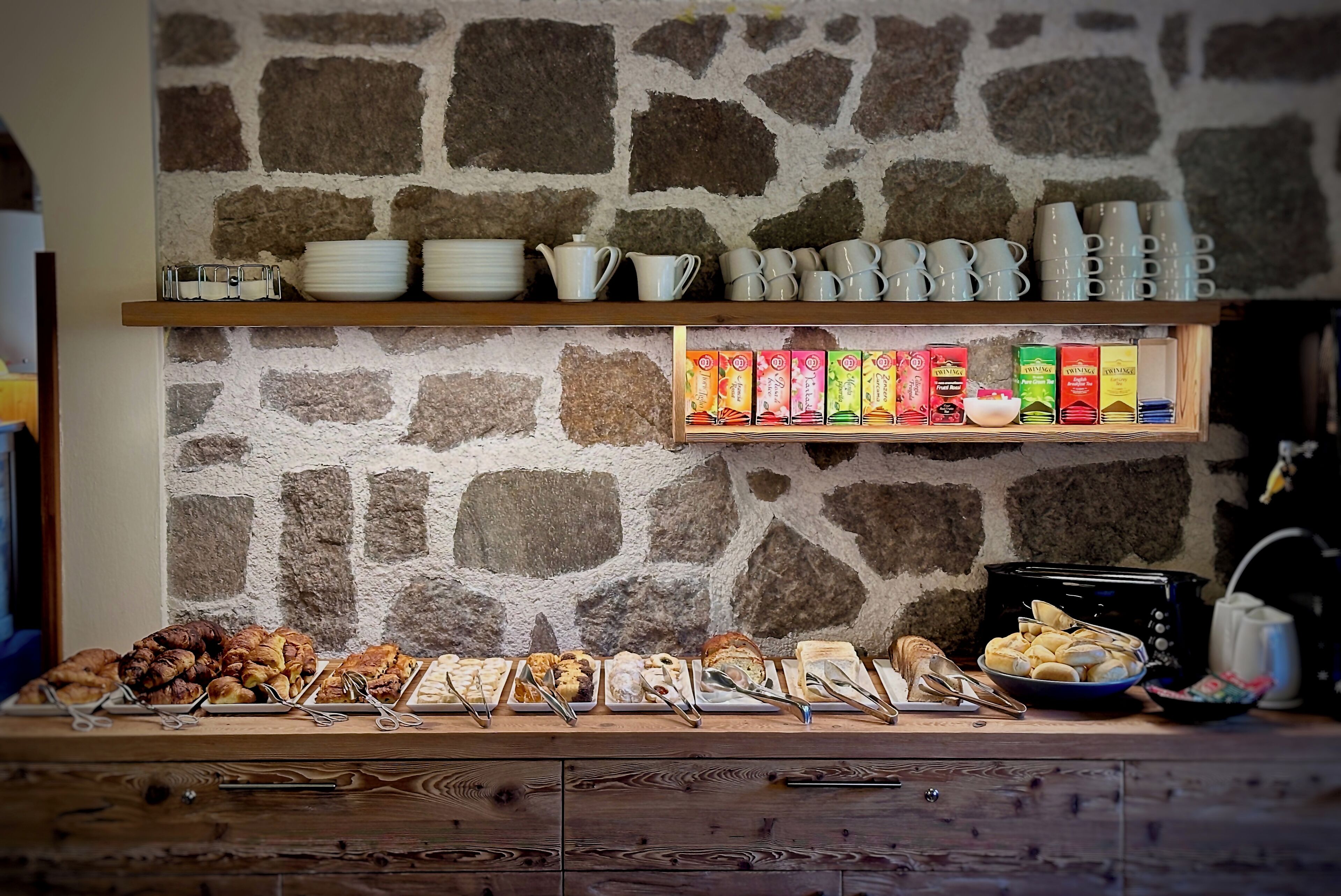 A buffet setup with various food items and beverages, including pastries, bread, and boxed drinks, against a stone wall backdrop.