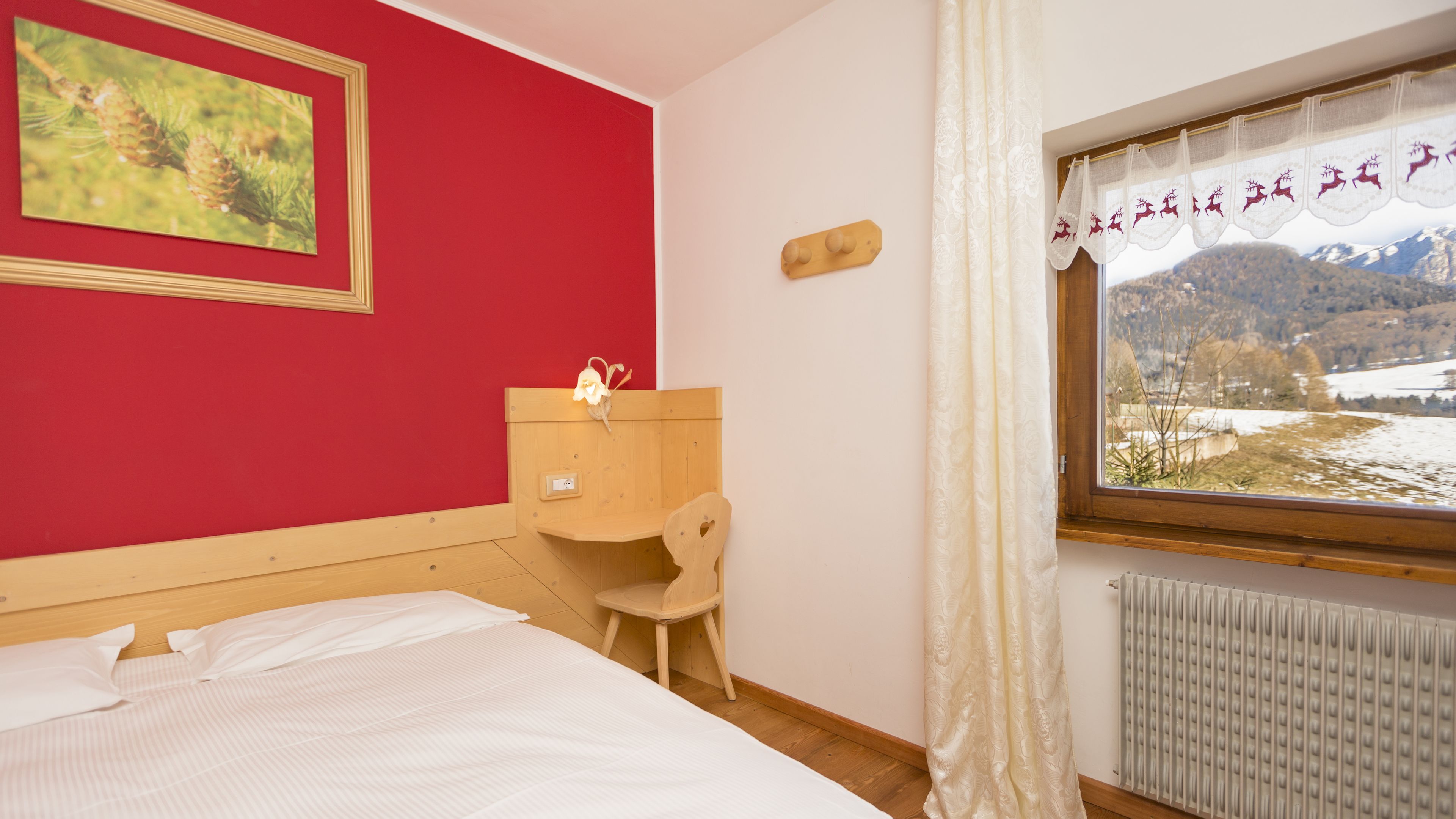 A bedroom with a bed, a wooden nightstand, a chair, a window with a view of a snowy landscape, and a red wall with a framed picture.